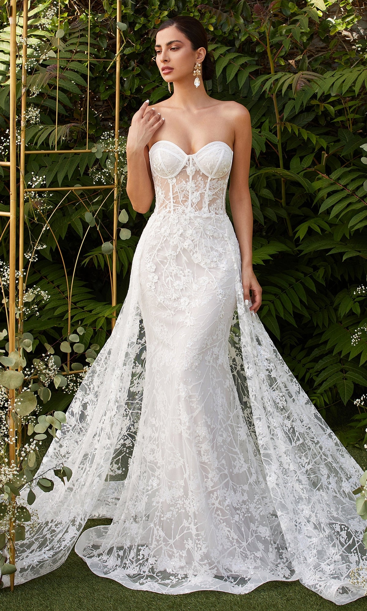A woman stands outdoors among lush green foliage wearing the Strapless Long White Wedding Dress CB046W, featuring delicate floral embroidery, a fitted sheer bodice, and a flowing train.